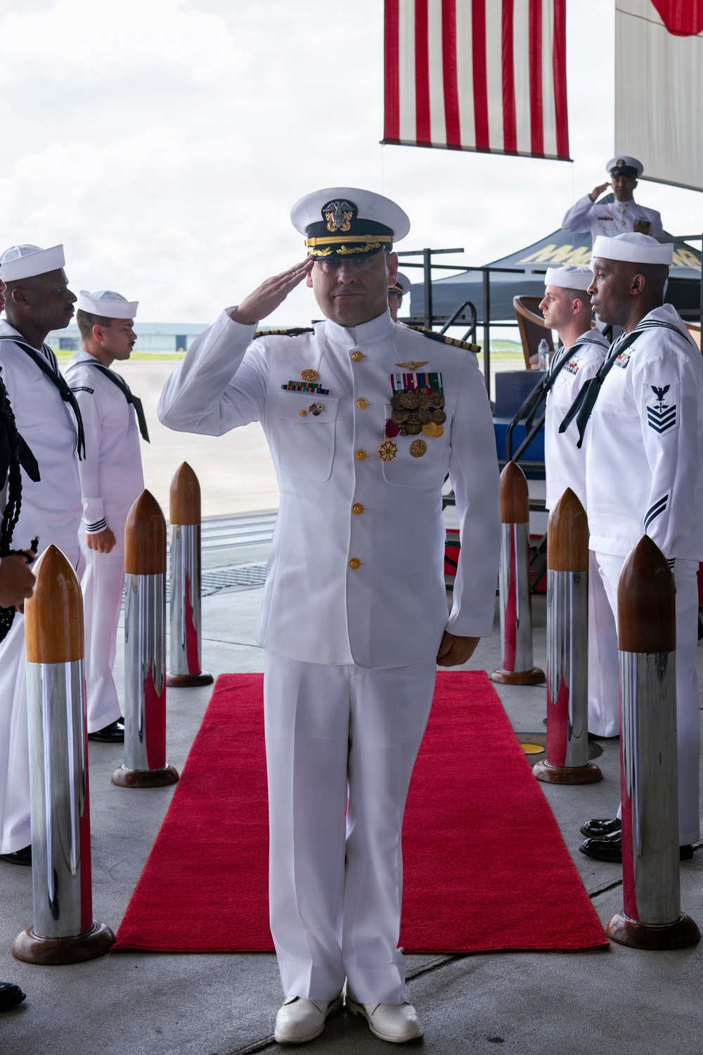 A U.S. Navy officer in white dress uniform is rendering a salute while standing on a red carpet flanked by ceremonial stanchions with decorative rope posts. The scene appears to be a formal military ceremony, likely a change of command or official arrival/departure event. An American flag is visible in the background along with additional Navy personnel in white uniforms forming a sideboy honor guard.