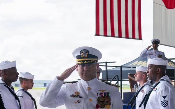 A U.S. Navy officer in white dress uniform is rendering a salute while standing on a red carpet flanked by ceremonial stanchions with decorative rope posts. The scene appears to be a formal military ceremony, likely a change of command or official arrival/departure event. An American flag is visible in the background along with additional Navy personnel in white uniforms forming a sideboy honor guard.