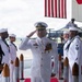 A U.S. Navy officer in white dress uniform is rendering a salute while standing on a red carpet flanked by ceremonial stanchions with decorative rope posts. The scene appears to be a formal military ceremony, likely a change of command or official arrival/departure event. An American flag is visible in the background along with additional Navy personnel in white uniforms forming a sideboy honor guard.