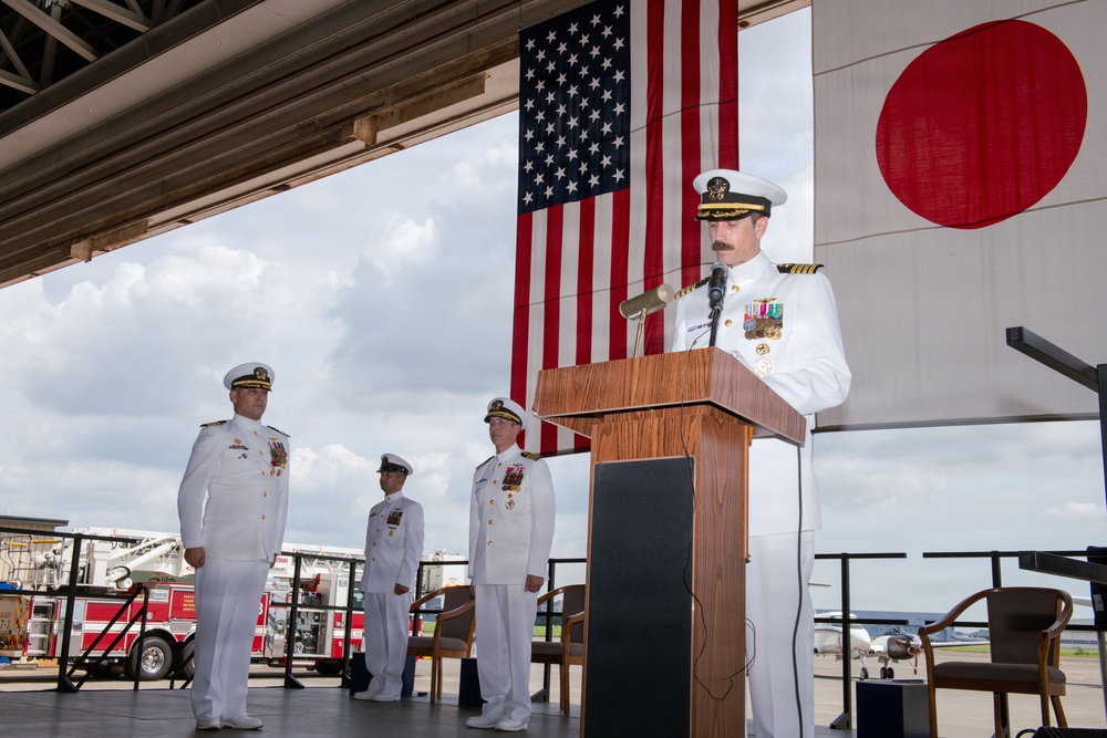 A formal U.S. Navy military ceremony is taking place in what appears to be an aircraft hangar or flight deck facility. A naval officer in white dress uniform is speaking at a wooden podium, flanked by the American flag and the Japanese flag, with three other uniformed personnel standing at attention in the background. The setting and dual flags suggest a bilateral military event, likely a change of command or official ceremony at a U.S. naval installation in Japan.