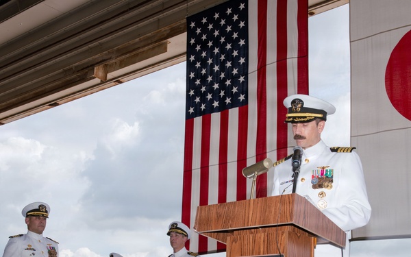 A formal U.S. Navy military ceremony is taking place in what appears to be an aircraft hangar or flight deck facility. A naval officer in white dress uniform is speaking at a wooden podium, flanked by the American flag and the Japanese flag, with three other uniformed personnel standing at attention in the background. The setting and dual flags suggest a bilateral military event, likely a change of command or official ceremony at a U.S. naval installation in Japan.