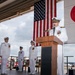 A formal U.S. Navy military ceremony is taking place in what appears to be an aircraft hangar or flight deck facility. A naval officer in white dress uniform is speaking at a wooden podium, flanked by the American flag and the Japanese flag, with three other uniformed personnel standing at attention in the background. The setting and dual flags suggest a bilateral military event, likely a change of command or official ceremony at a U.S. naval installation in Japan.