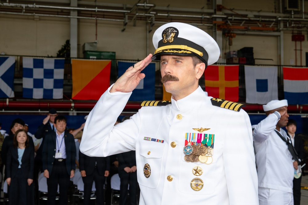 A U.S. Navy officer in white dress uniform is rendering a salute at what appears to be a formal military ceremony. The officer displays multiple medals, ribbons, and insignia consistent with a senior rank, likely a Captain or Commander. The event appears to be taking place in a hangar or covered facility decorated with international maritime signal flags.