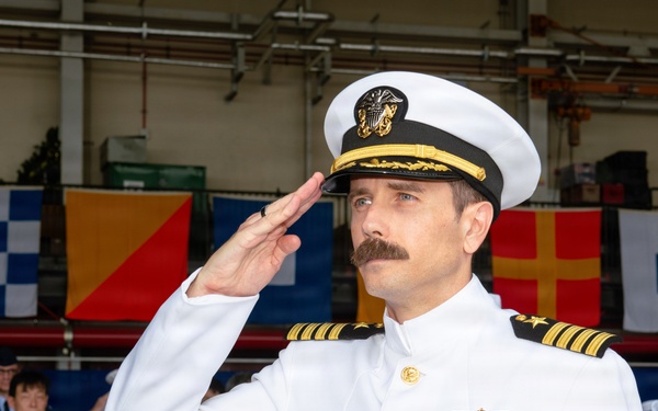 A U.S. Navy officer in white dress uniform is rendering a salute at what appears to be a formal military ceremony. The officer displays multiple medals, ribbons, and insignia consistent with a senior rank, likely a Captain or Commander. The event appears to be taking place in a hangar or covered facility decorated with international maritime signal flags.
