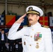 A U.S. Navy officer in white dress uniform is rendering a salute at what appears to be a formal military ceremony. The officer displays multiple medals, ribbons, and insignia consistent with a senior rank, likely a Captain or Commander. The event appears to be taking place in a hangar or covered facility decorated with international maritime signal flags.
