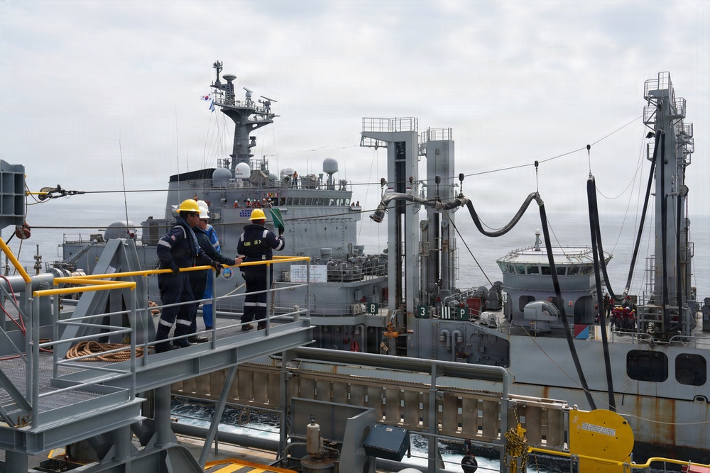 A replenishment-at-sea (RAS) operation is underway between multiple naval vessels on open ocean. Two crew members wearing yellow hard hats and blue coveralls are visible on the foreground vessel's deck, monitoring the operation. Transfer hoses and rigging lines are visibly connected between the ships.