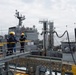 A replenishment-at-sea (RAS) operation is underway between multiple naval vessels on open ocean. Two crew members wearing yellow hard hats and blue coveralls are visible on the foreground vessel's deck, monitoring the operation. Transfer hoses and rigging lines are visibly connected between the ships.