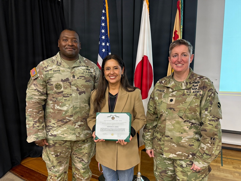 A civilian woman named Veronica Lewis is being recognized with a Department of the Army Civilian Service Commendation Medal certificate. She is flanked by two U.S. Army soldiers — one identified by nametape as 'Davis' on the left and 'Johnson' on the right — in a formal award ceremony setting. The event appears to take place in Japan, as indicated by the Japanese flag visible in the background.