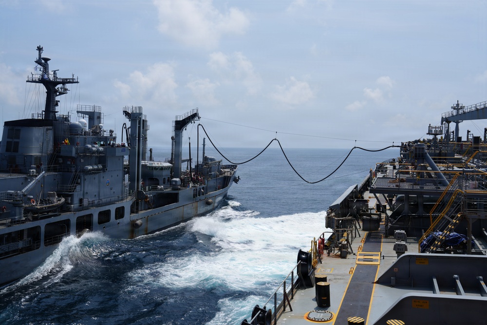 The image depicts a Replenishment at Sea (RAS) operation between two naval vessels underway in open ocean conditions. Two hoses or transfer lines are visibly strung between the ships, indicating an active fuel or liquid transfer operation. The ships are sailing in close parallel formation with visible wake turbulence between them.