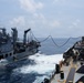 The image depicts a Replenishment at Sea (RAS) operation between two naval vessels underway in open ocean conditions. Two hoses or transfer lines are visibly strung between the ships, indicating an active fuel or liquid transfer operation. The ships are sailing in close parallel formation with visible wake turbulence between them.