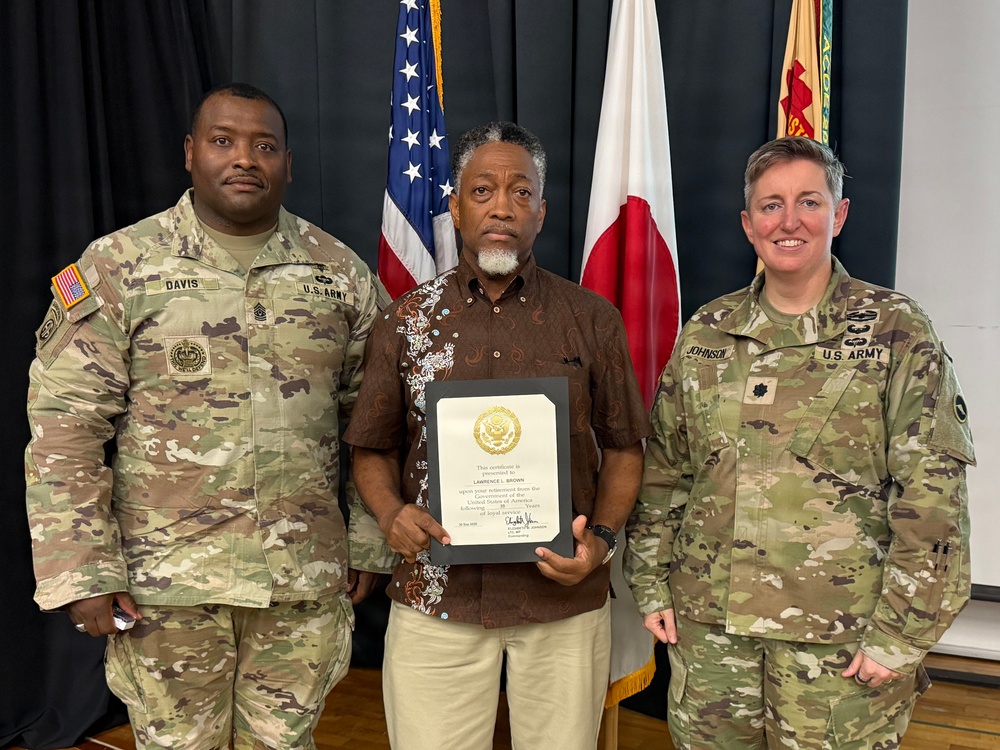 Three individuals pose for a formal photo at what appears to be a U.S. Army ceremony, likely held in Japan based on the visible Japanese flag. The central civilian figure, identified on the certificate as Lawrence L. Brown, holds a framed certificate of recognition presented by the U.S. Government. Two U.S. Army soldiers in MultiCam (OCP) uniforms flank him, identified by name tapes as Davis (left) and Johnson (right).