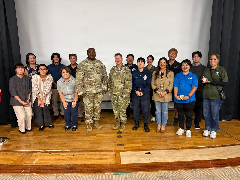 A group of approximately 17 people pose for a formal group photo on a stage or auditorium platform in front of a white projection screen. The group includes two individuals in U.S. Army camouflage (OCP) uniforms, one person in a law enforcement or security uniform, and several civilians in business casual and casual attire. Some individuals appear to be wearing organizational polo shirts, suggesting a multi-agency or community event.