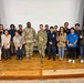 A group of approximately 17 people pose for a formal group photo on a stage or auditorium platform in front of a white projection screen. The group includes two individuals in U.S. Army camouflage (OCP) uniforms, one person in a law enforcement or security uniform, and several civilians in business casual and casual attire. Some individuals appear to be wearing organizational polo shirts, suggesting a multi-agency or community event.