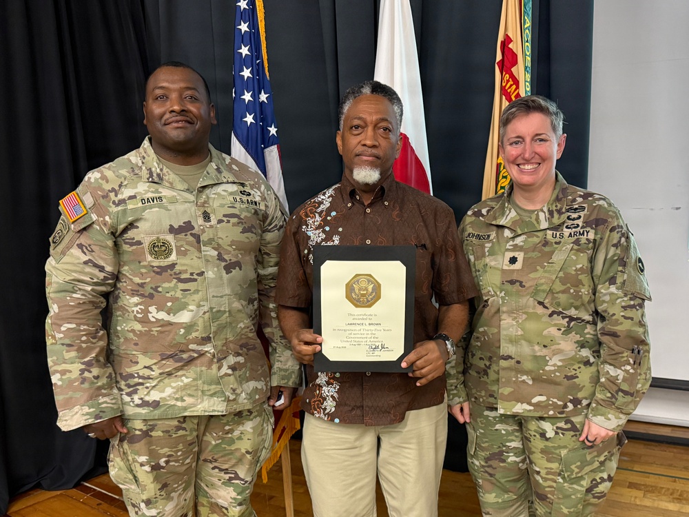 Three individuals pose for a formal photo at what appears to be a U.S. Army recognition ceremony. The civilian in the center, identified as Lawrence L. Brown on the certificate he holds, is being honored for Thirty-Five Years of service to the United States Army. He is flanked by two U.S. Army soldiers in MultiCam uniforms, identified by name tapes as Davis (left) and Johnson (right).