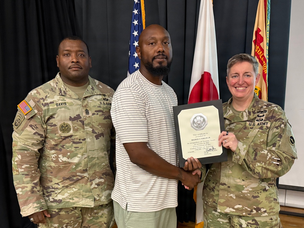 Three individuals pose for a formal recognition photo, with a civilian man in the center holding a certificate alongside a U.S. Army female soldier on the right and a male U.S. Army soldier named Davis on the left. The certificate appears to be awarded to Maurice V. Fowler Jr. in recognition of twenty years of service to the United States Government. The setting appears to be an official military ceremony room with American and other flags visible in the background.