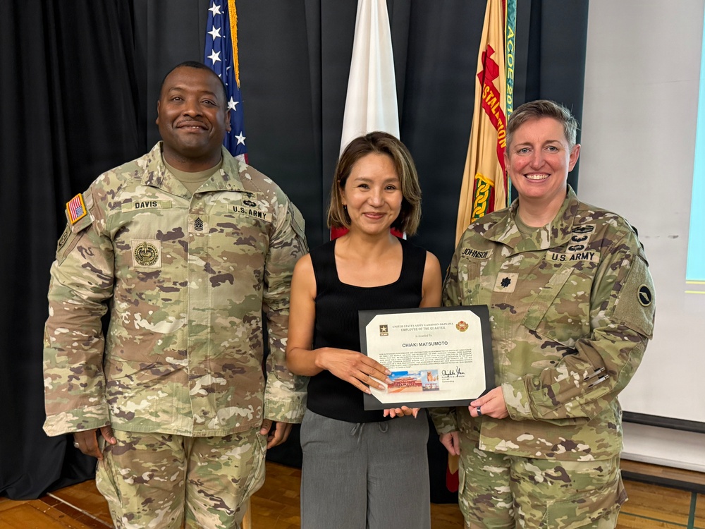 Three individuals pose for a formal recognition photo at what appears to be a U.S. Army garrison ceremony. The civilian in the center, identified as Chiaki Matsumoto per the certificate, is being recognized as 'Employee of the Quarter' by two U.S. Army personnel flanking her. The setting includes multiple flags including the U.S. flag and a Japanese flag in the background.