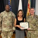 Three individuals pose for a formal recognition photo at what appears to be a U.S. Army garrison ceremony. The civilian in the center, identified as Chiaki Matsumoto per the certificate, is being recognized as 'Employee of the Quarter' by two U.S. Army personnel flanking her. The setting includes multiple flags including the U.S. flag and a Japanese flag in the background.