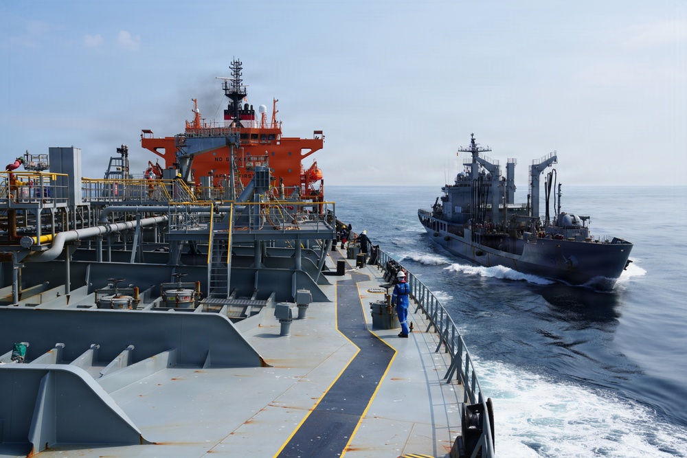 The image shows two vessels at sea, with a large tanker ship featuring a distinctive orange superstructure in the foreground and a gray naval or auxiliary vessel approaching from the starboard side. A crew member in blue coveralls and a white hard hat is visible walking along the deck of the foreground vessel. The scene appears to depict a replenishment-at-sea (RAS) or ship-to-ship transfer operation in open ocean conditions.