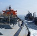 The image shows two vessels at sea, with a large tanker ship featuring a distinctive orange superstructure in the foreground and a gray naval or auxiliary vessel approaching from the starboard side. A crew member in blue coveralls and a white hard hat is visible walking along the deck of the foreground vessel. The scene appears to depict a replenishment-at-sea (RAS) or ship-to-ship transfer operation in open ocean conditions.