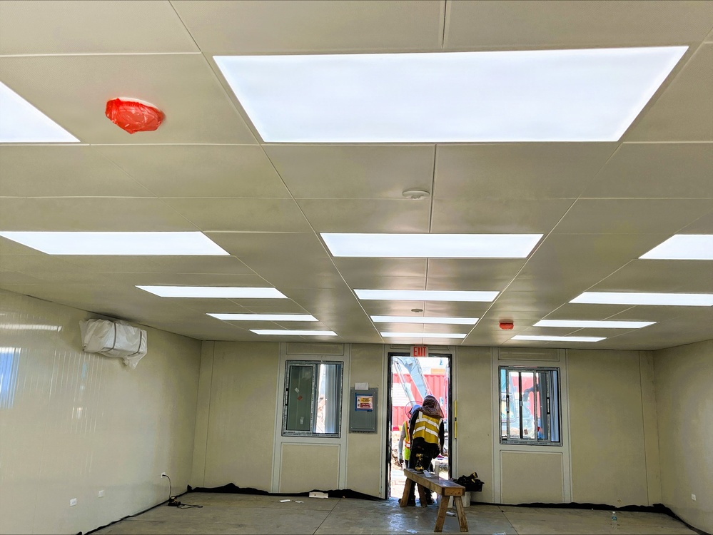 The image shows the interior of a modular or prefabricated building under construction or fit-out, featuring a drop ceiling with large LED panel lights installed throughout. A worker in a high-visibility vest is standing near the open doorway at the far end, through which construction equipment and red shipping containers are visible outside. The space appears to be a temporary site office or welfare facility in the process of being completed.