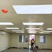 The image shows the interior of a modular or prefabricated building under construction or fit-out, featuring a drop ceiling with large LED panel lights installed throughout. A worker in a high-visibility vest is standing near the open doorway at the far end, through which construction equipment and red shipping containers are visible outside. The space appears to be a temporary site office or welfare facility in the process of being completed.