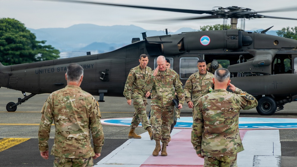 A group of U.S. Army personnel in multicam/OCP uniforms are conducting a formal arrival ceremony on a helicopter landing pad. A senior officer is walking down a red carpet pathway from a UH-60 Black Hawk helicopter marked 'UNITED STATES ARMY,' while two soldiers render salutes. The setting appears to be an overseas military installation, likely in a tropical region based on the vegetation and mountainous background.