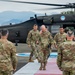 A group of U.S. Army personnel in multicam/OCP uniforms are conducting a formal arrival ceremony on a helicopter landing pad. A senior officer is walking down a red carpet pathway from a UH-60 Black Hawk helicopter marked 'UNITED STATES ARMY,' while two soldiers render salutes. The setting appears to be an overseas military installation, likely in a tropical region based on the vegetation and mountainous background.