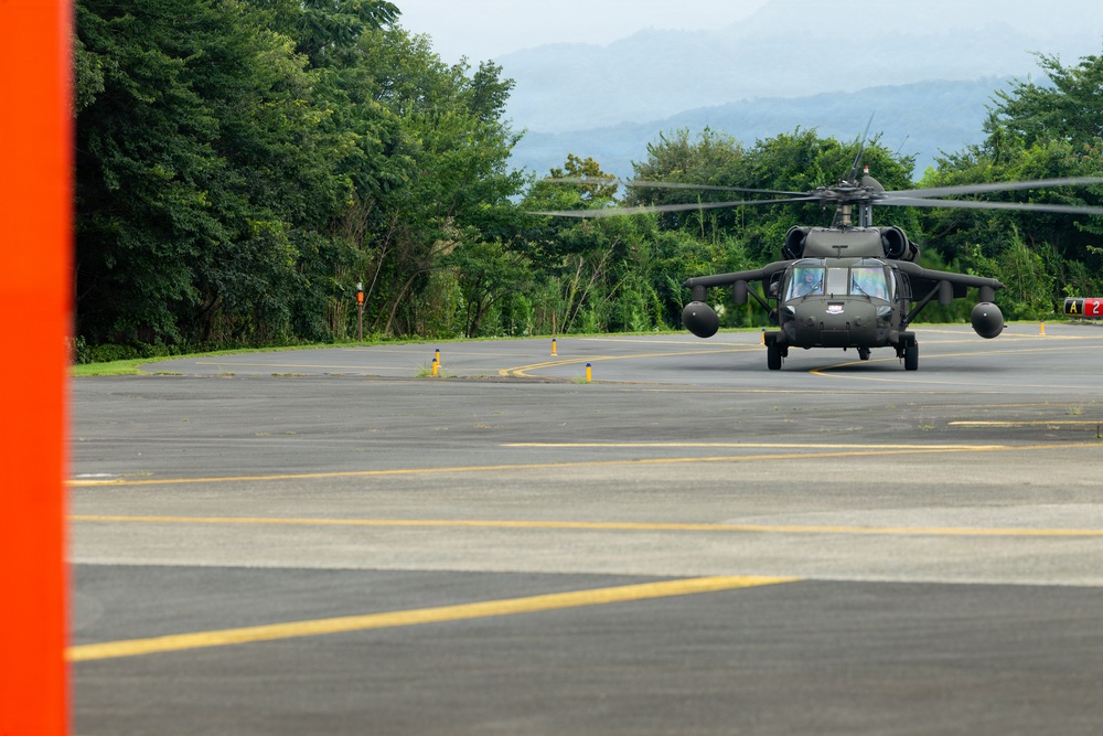 A military helicopter, consistent with a UH-60 Black Hawk, is positioned on a paved tarmac or helipad area. The aircraft appears to be either taxiing or stationary with rotors spinning, set against a backdrop of dense green trees and distant mountains. The setting suggests a military airfield or forward operating base in a tropical or subtropical region.