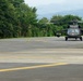 A military helicopter, consistent with a UH-60 Black Hawk, is positioned on a paved tarmac or helipad area. The aircraft appears to be either taxiing or stationary with rotors spinning, set against a backdrop of dense green trees and distant mountains. The setting suggests a military airfield or forward operating base in a tropical or subtropical region.