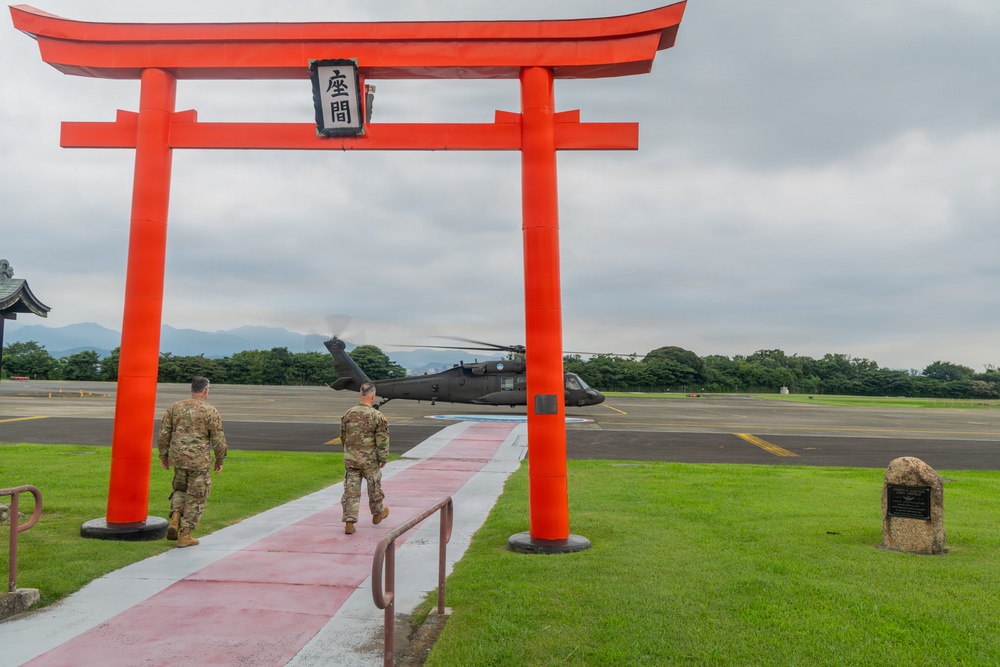 Two U.S. Army soldiers in camouflage uniforms walk through a large red torii gate toward a military helicopter on a tarmac. The torii gate bears the kanji characters '座間' (Zama), indicating this is Camp Zama, a U.S. Army installation in Japan. A UH-60 Black Hawk helicopter is visible on the flight line in the background.