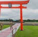 Two U.S. Army soldiers in camouflage uniforms walk through a large red torii gate toward a military helicopter on a tarmac. The torii gate bears the kanji characters '座間' (Zama), indicating this is Camp Zama, a U.S. Army installation in Japan. A UH-60 Black Hawk helicopter is visible on the flight line in the background.