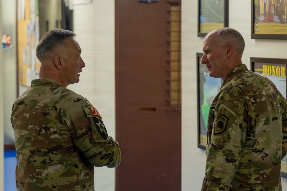 Two U.S. Army soldiers in Operational Camouflage Pattern (OCP) uniforms are engaged in conversation inside what appears to be a military facility. The soldier on the left has his arms crossed and displays a U.S. flag patch and a unit patch on his left sleeve. They are standing in a hallway or common area with motivational military posters visible on the wall in the background.