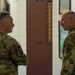 Two U.S. Army soldiers in Operational Camouflage Pattern (OCP) uniforms are engaged in conversation inside what appears to be a military facility. The soldier on the left has his arms crossed and displays a U.S. flag patch and a unit patch on his left sleeve. They are standing in a hallway or common area with motivational military posters visible on the wall in the background.