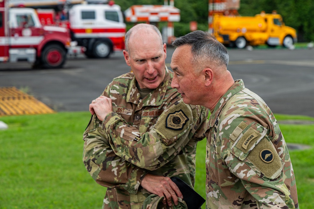 Two military personnel in OCP (Operational Camouflage Pattern) uniforms are engaged in close conversation outdoors. One individual wears a US Air Force uniform with a command patch, while the other displays a Ranger tab and what appears to be an Army unit patch. They are standing on a paved area with emergency and utility vehicles visible in the background.