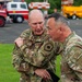 Two military personnel in OCP (Operational Camouflage Pattern) uniforms are engaged in close conversation outdoors. One individual wears a US Air Force uniform with a command patch, while the other displays a Ranger tab and what appears to be an Army unit patch. They are standing on a paved area with emergency and utility vehicles visible in the background.