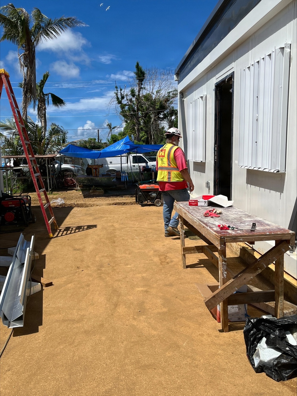 A construction worker wearing a yellow high-visibility vest, red shirt, and white hard hat is walking alongside a prefabricated or modular metal building at an active construction site. The surrounding area shows signs of storm or hurricane damage, with stripped and damaged palm trees visible in the background. Multiple generators, a ladder, a work table with tools, and a blue tarp canopy indicate ongoing recovery or rebuilding operations.