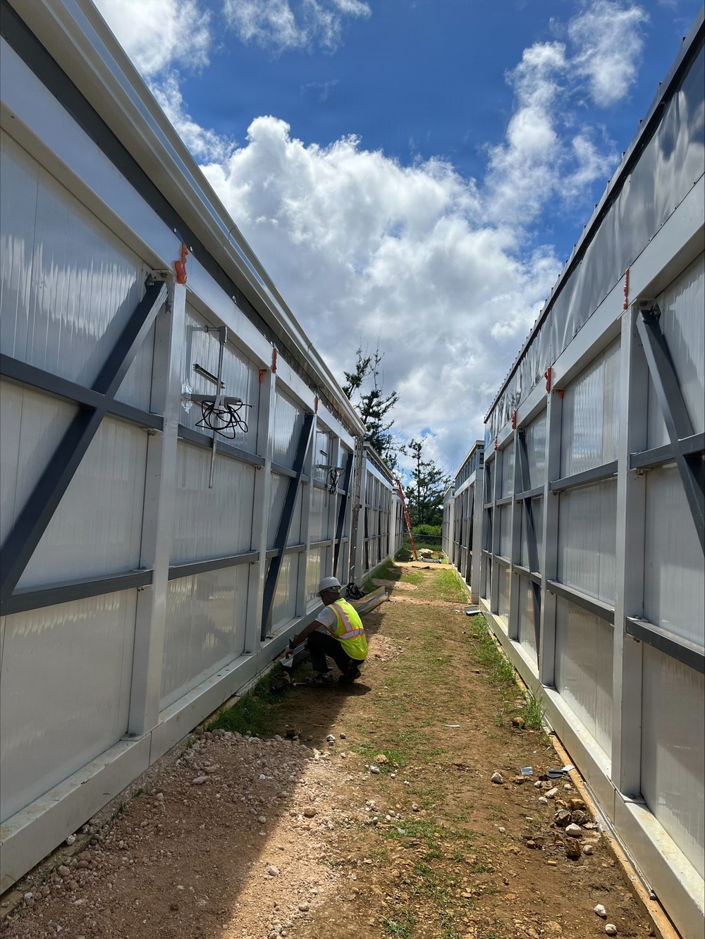 A construction worker in high-visibility safety gear and hard hat is crouching at the base of a structure between two parallel rows of modular or prefabricated panel walls with translucent polycarbonate sections and gray steel framing. The narrow corridor between the two structures leads toward a tree line and blue sky in the background. The site appears to be an active construction zone with loose soil, rocks, and scattered materials on the ground.