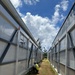 A construction worker in high-visibility safety gear and hard hat is crouching at the base of a structure between two parallel rows of modular or prefabricated panel walls with translucent polycarbonate sections and gray steel framing. The narrow corridor between the two structures leads toward a tree line and blue sky in the background. The site appears to be an active construction zone with loose soil, rocks, and scattered materials on the ground.