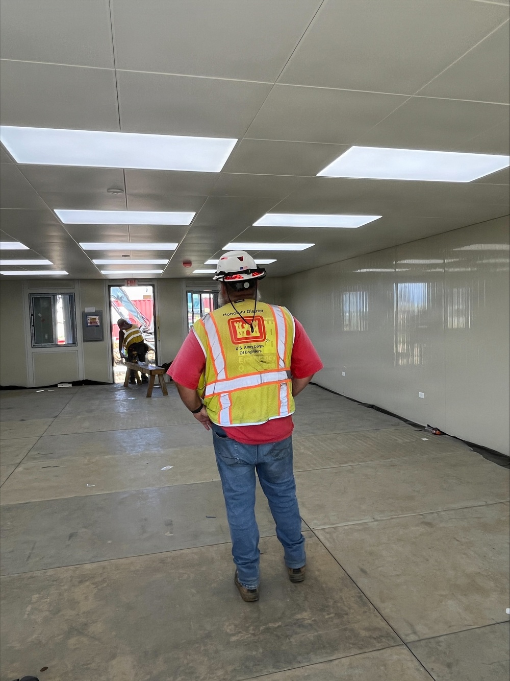 A U.S. Army Corps of Engineers inspector from the Honolulu District is conducting an interior inspection of a modular or prefabricated building under construction. The individual is wearing a high-visibility safety vest with the USACE castle logo and 'Honolulu District' text, along with a white hard hat. The interior space appears to be in the final stages of construction with installed ceiling panels, LED lighting, and smooth wall panels.