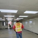 A U.S. Army Corps of Engineers inspector from the Honolulu District is conducting an interior inspection of a modular or prefabricated building under construction. The individual is wearing a high-visibility safety vest with the USACE castle logo and 'Honolulu District' text, along with a white hard hat. The interior space appears to be in the final stages of construction with installed ceiling panels, LED lighting, and smooth wall panels.