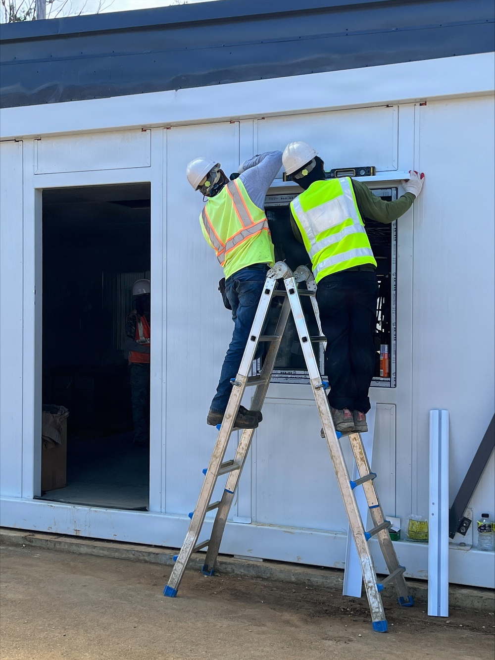 Two construction workers in high-visibility safety vests and hard hats are standing on a shared aluminum ladder, working on what appears to be an electrical panel or equipment installation on the exterior wall of a white modular or prefabricated building. One worker is using a level to ensure proper alignment of the panel. A third worker in safety gear is visible in the background through an open doorway.