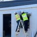Two construction workers in high-visibility safety vests and hard hats are standing on a shared aluminum ladder, working on what appears to be an electrical panel or equipment installation on the exterior wall of a white modular or prefabricated building. One worker is using a level to ensure proper alignment of the panel. A third worker in safety gear is visible in the background through an open doorway.