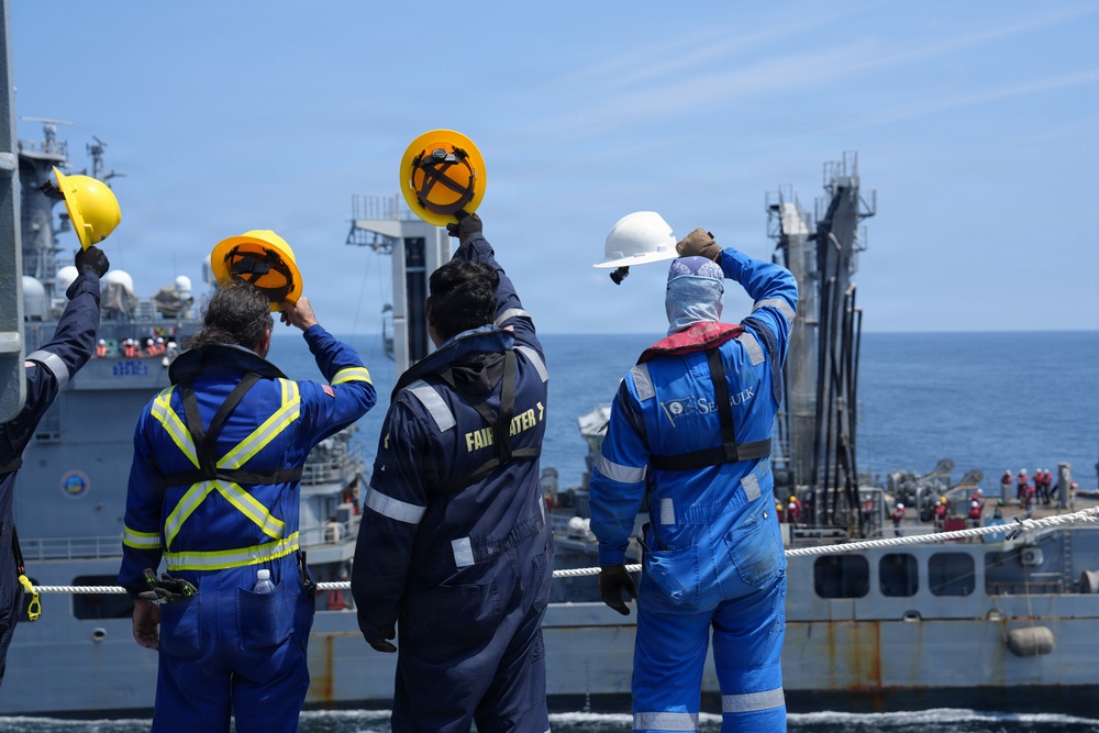 Four maritime workers in safety gear are standing on the deck of a vessel, raising their hard hats in a farewell or celebratory gesture toward another ship nearby. The workers are wearing blue coveralls and high-visibility vests, suggesting a professional maritime or naval replenishment operation. A second large vessel is visible in the background with additional crew members on its deck.
