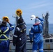 Four maritime workers in safety gear are standing on the deck of a vessel, raising their hard hats in a farewell or celebratory gesture toward another ship nearby. The workers are wearing blue coveralls and high-visibility vests, suggesting a professional maritime or naval replenishment operation. A second large vessel is visible in the background with additional crew members on its deck.