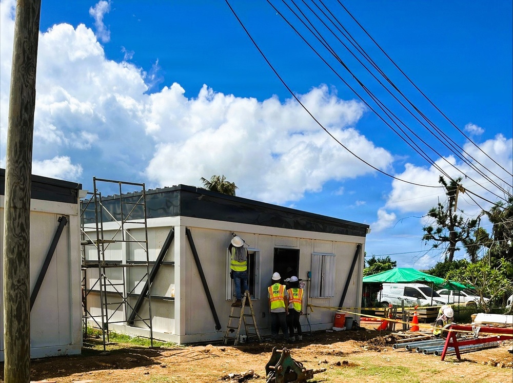 A construction or renovation site showing workers in high-visibility safety vests and hard hats working on a modular or prefabricated building. The structure features distinctive black diagonal bracing on its exterior walls and a flat dark roof. The surrounding environment shows signs of storm or hurricane damage, including stripped and damaged trees.