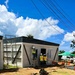 A construction or renovation site showing workers in high-visibility safety vests and hard hats working on a modular or prefabricated building. The structure features distinctive black diagonal bracing on its exterior walls and a flat dark roof. The surrounding environment shows signs of storm or hurricane damage, including stripped and damaged trees.