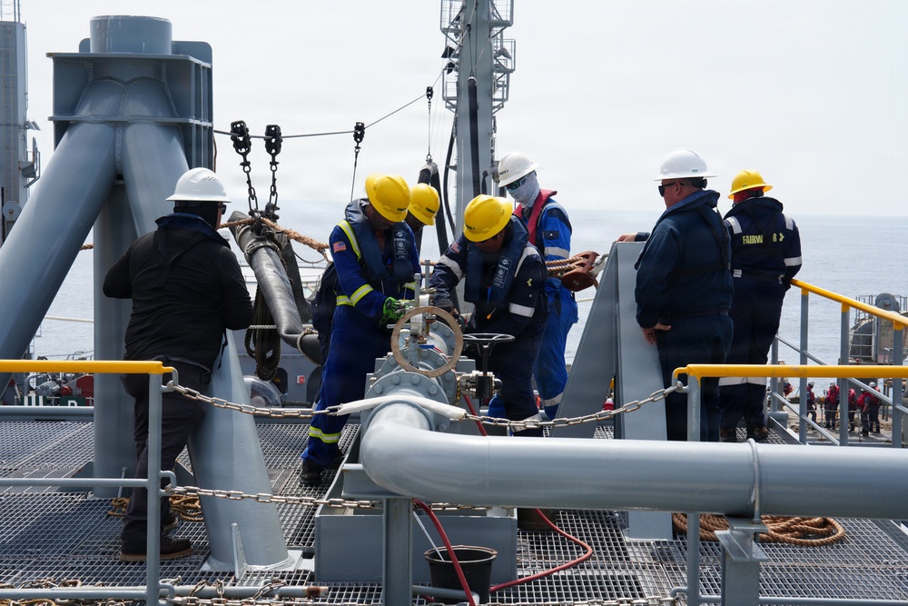 A group of workers in personal protective equipment are performing operations on the deck of a naval or industrial vessel at sea. They appear to be handling a valve or pipe connection, with some personnel managing ropes and others observing. The scene suggests a replenishment-at-sea (RAS) or similar maritime industrial operation.