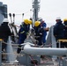A group of workers in personal protective equipment are performing operations on the deck of a naval or industrial vessel at sea. They appear to be handling a valve or pipe connection, with some personnel managing ropes and others observing. The scene suggests a replenishment-at-sea (RAS) or similar maritime industrial operation.