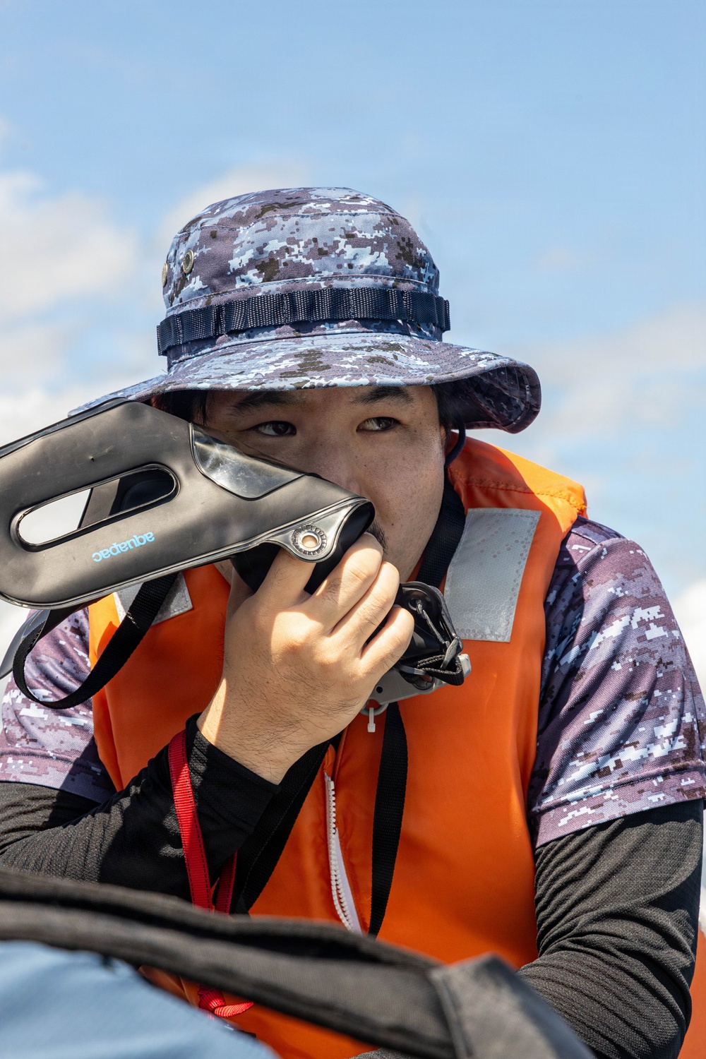 A person wearing an orange life vest and digital camouflage boonie hat holds an Aquapac waterproof case or device up to their face. The individual appears to be on or near a watercraft, suggesting a maritime or water-based activity. The image is a close-up portrait taken outdoors under a partly cloudy sky.