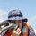 U.S. Marines, JMSDF Sailors reinforce maritime readiness during Rescue Flag 26-1 A person wearing an orange life vest and digital camouflage boonie hat holds an Aquapac waterproof case or device up to their face. The individual appears to be on or near a watercraft, suggesting a maritime or water-based activity. The image is a close-up portrait taken outdoors under a partly cloudy sky.